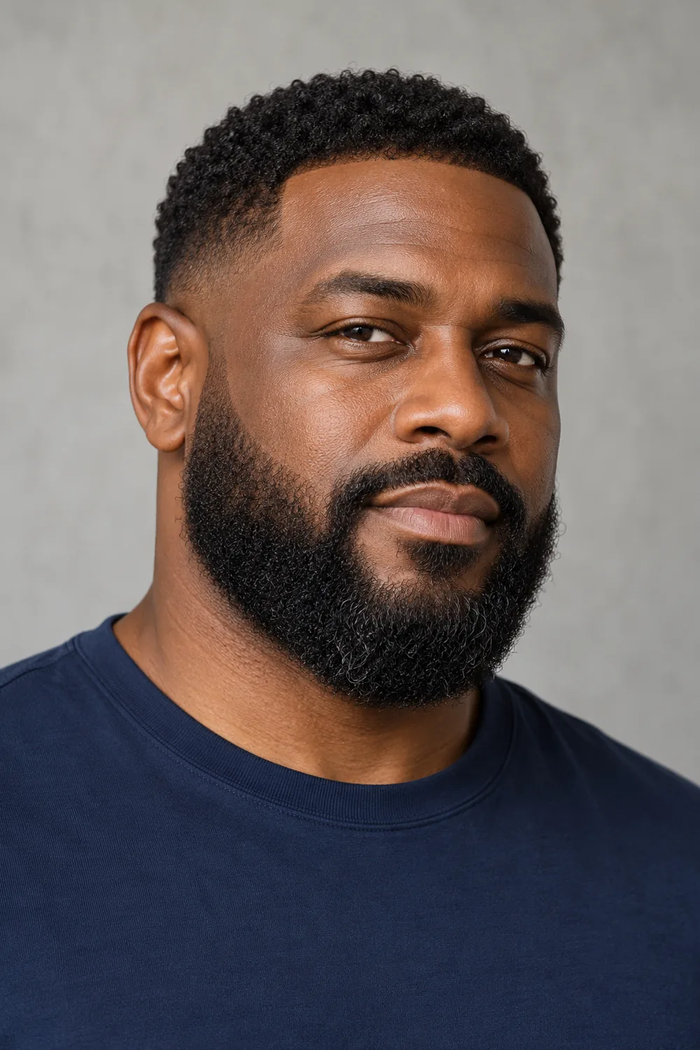 A fictional adult Black man in a navy crew-neck shirt, shown in three-quarter view with a dense rounded beard, connected mustache, soft cheek line, and low sideburn fade.