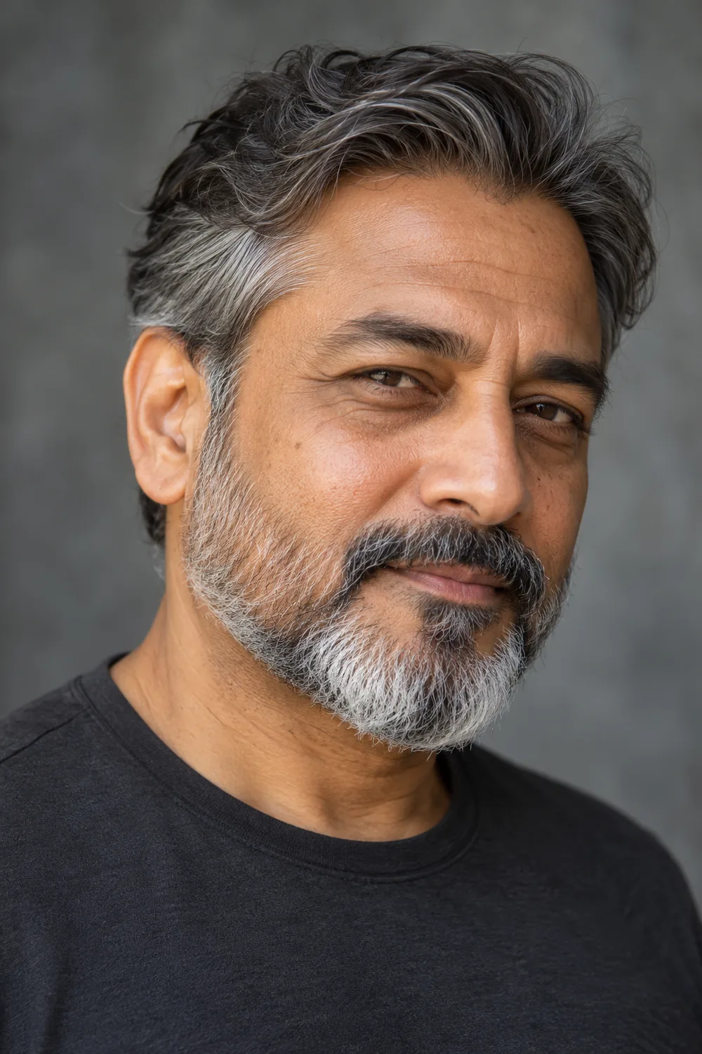 Mature South Asian man in a charcoal shirt with a close salt-and-pepper beard, connected mustache, and softly squared chin.