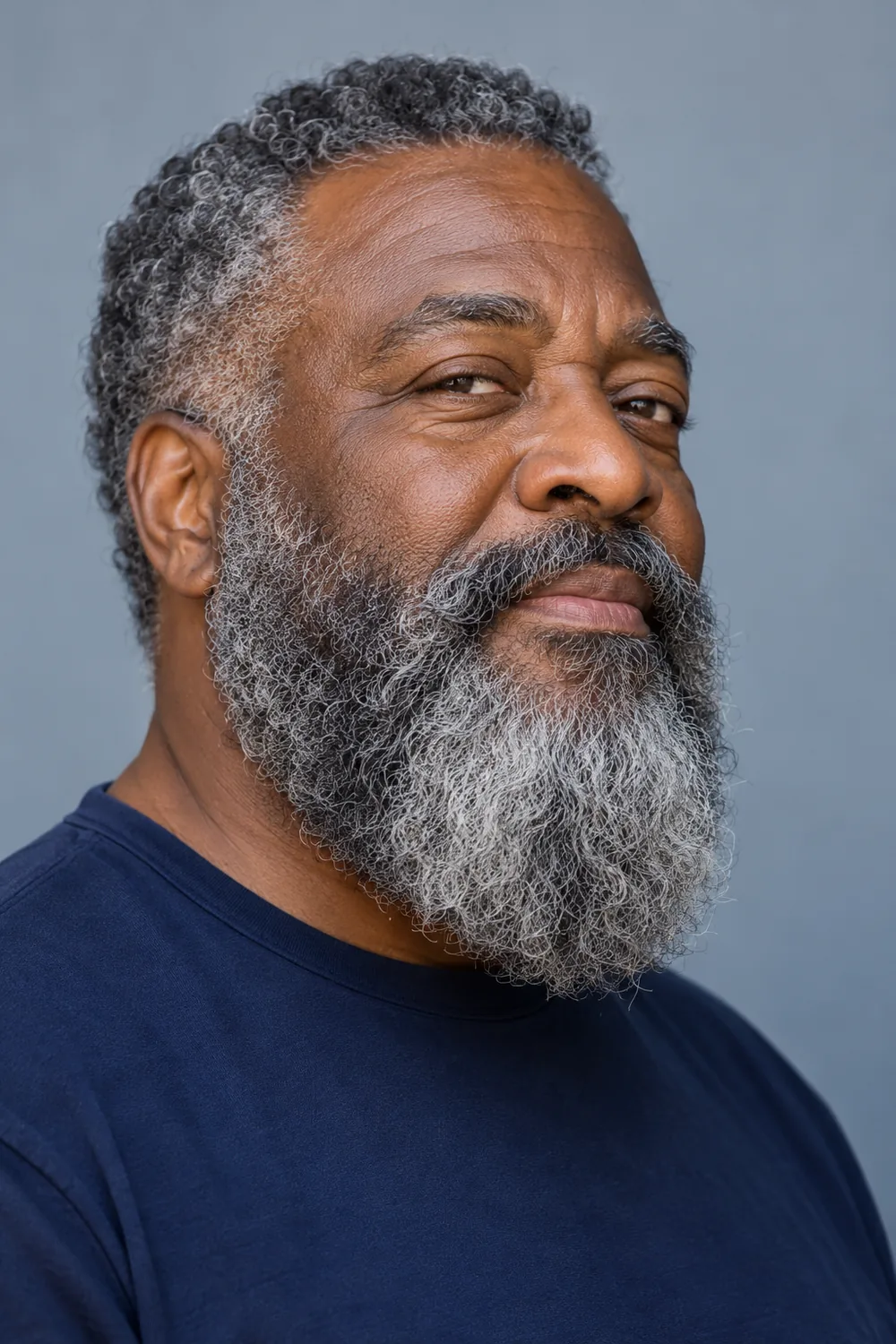 Three-quarter portrait of a mature Black man wearing a dense, rounded beard with tightly curled charcoal, silver, and white strands and a connected mustache.