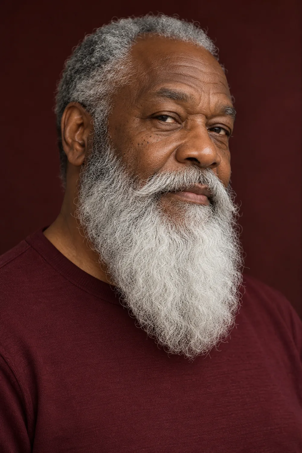 Older Black man with a long, mostly white wavy beard, low cheek line, and full connected mustache, wearing a burgundy crew-neck shirt.