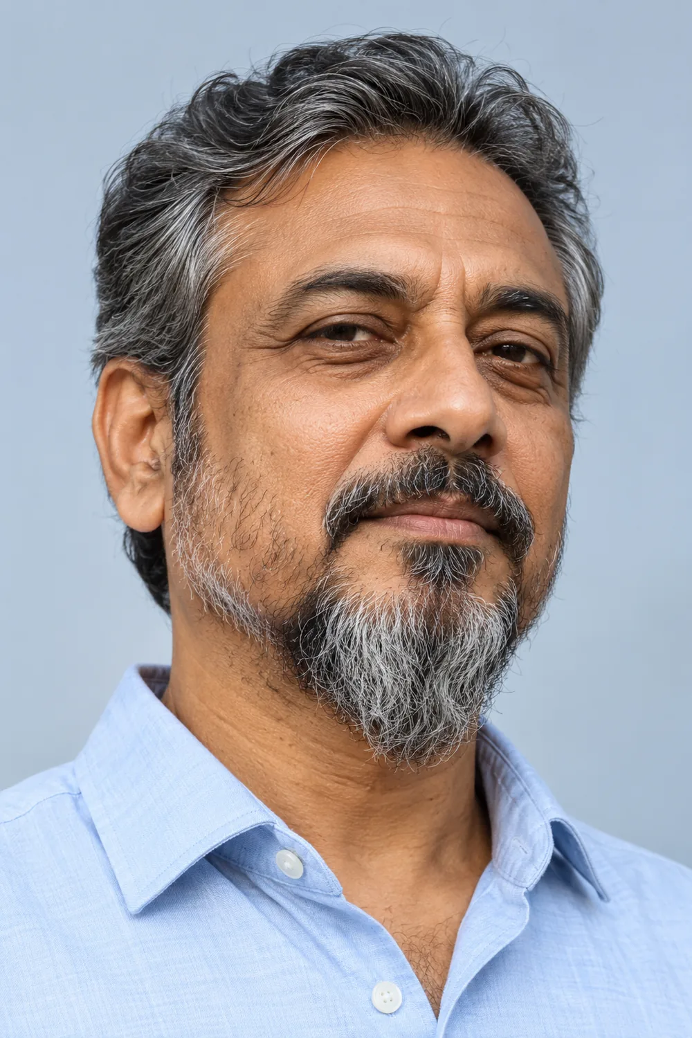 Three-quarter portrait of an adult South Asian man with very sparse cheek growth, a longer salt-and-pepper chin beard, and a modest mustache separated at the corners.
