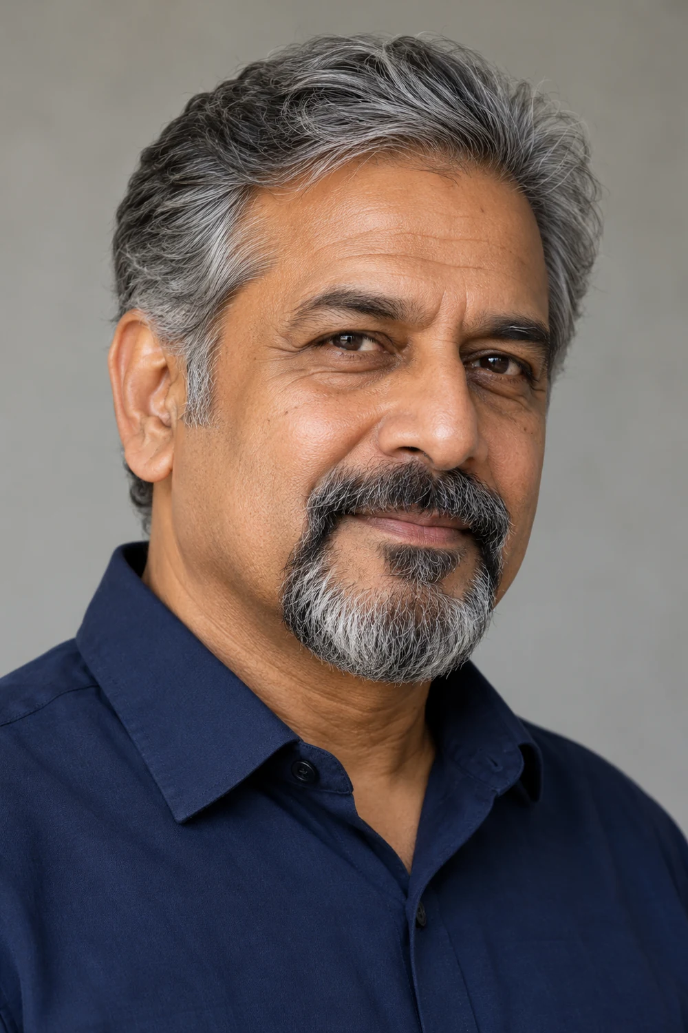 Adult South Asian man with salt-and-pepper hair and a medium-full circle beard, rounded chin and connected mustache, wearing a navy shirt.