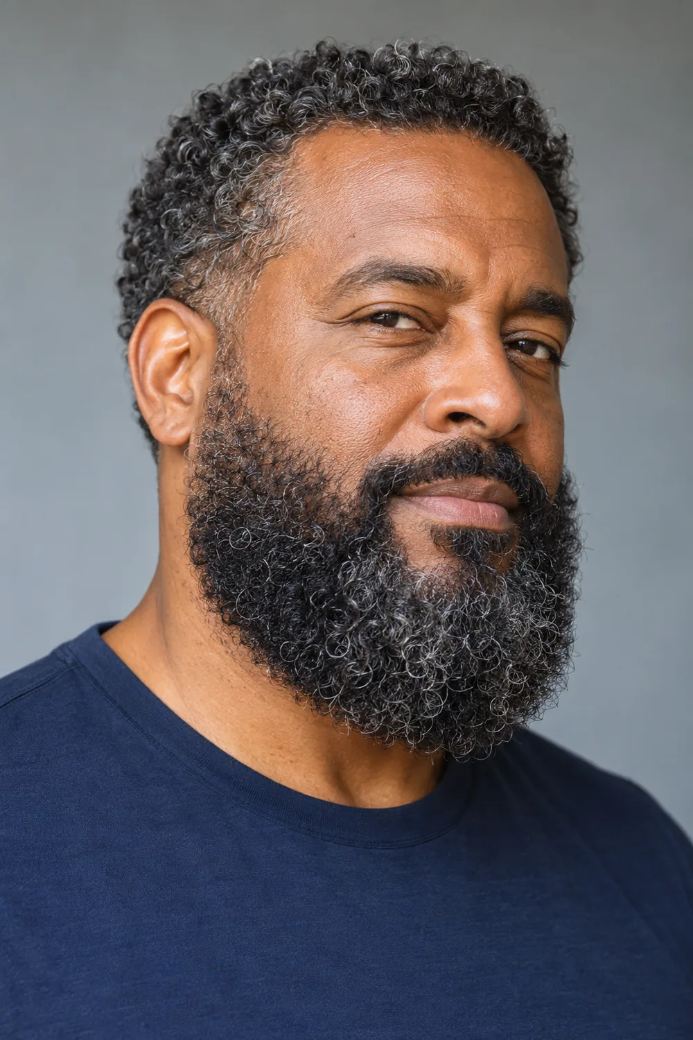 Three-quarter portrait of a middle-aged Black man with a dense, medium-length curly beard, low natural cheek line, rounded chin, and connected mustache.