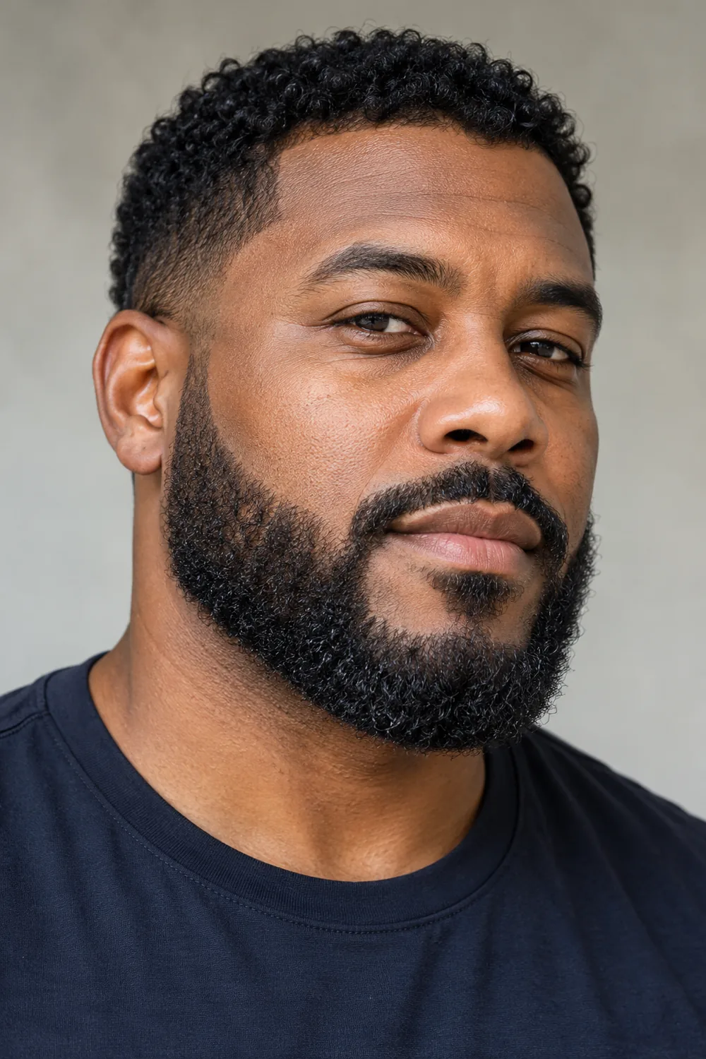 Three-quarter portrait of an adult Black man with dense coiled hair, a short boxed curly beard, crisp cheek and jaw edges, and a compact connected mustache.