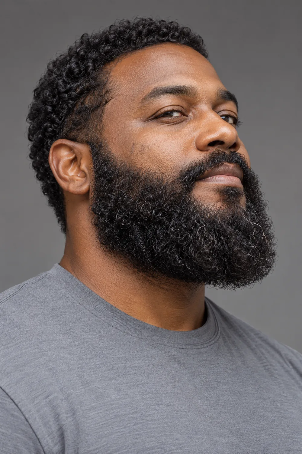 Upward three-quarter portrait of a man in a gray shirt with a full, dense curly beard, rounded chin, natural neckline, and blended mustache.