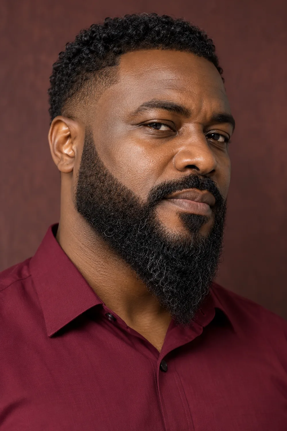 Adult man in a burgundy shirt with a dense, tightly curled beard, crisp high cheek lines, and a pronounced tapered chin point.