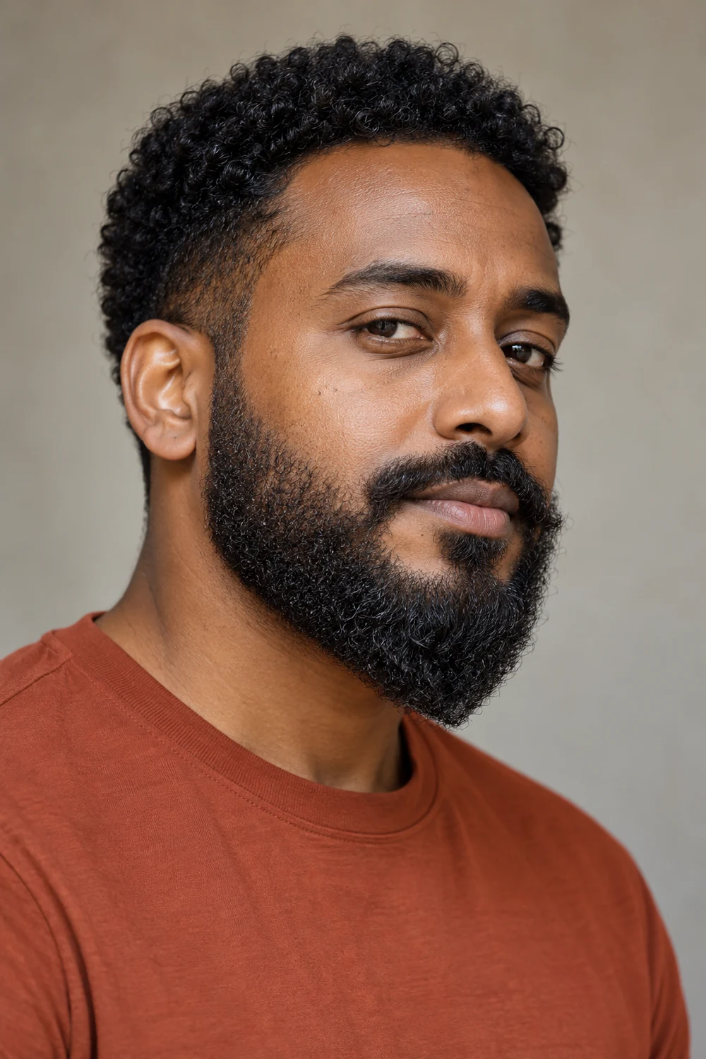 Adult man with a dense curly beard tapering to a compact, softly rounded ducktail point.