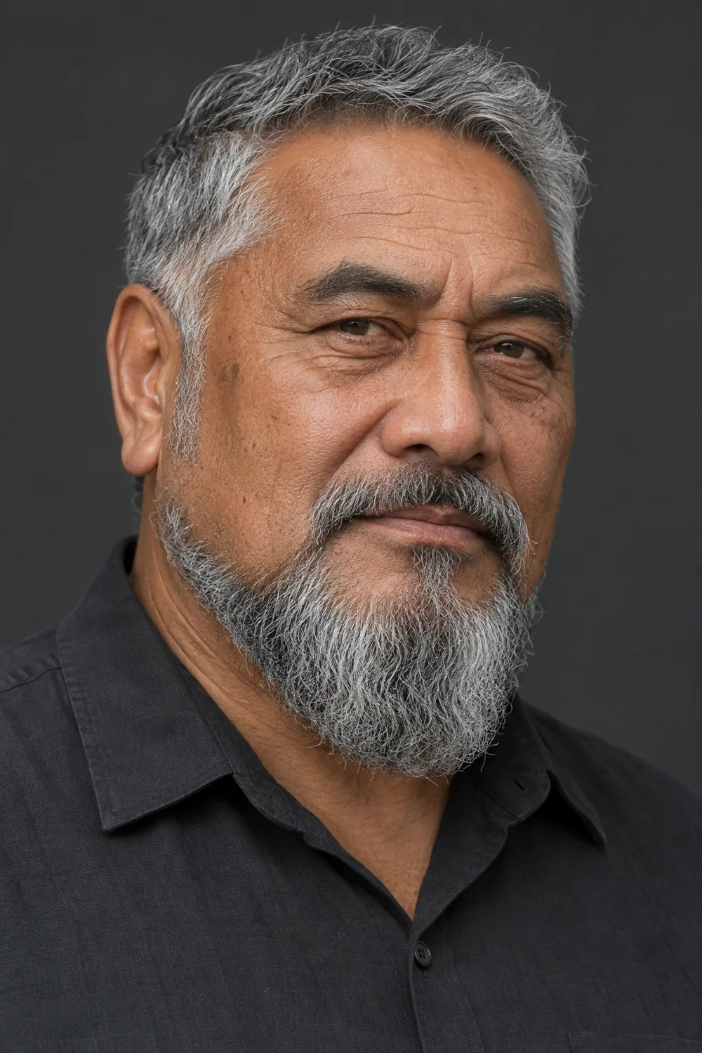 Three-quarter portrait of an older Māori man with a natural-textured gray extended goatee, rounded chin, connected mustache, shaved upper cheeks, and short disconnected sideburns.