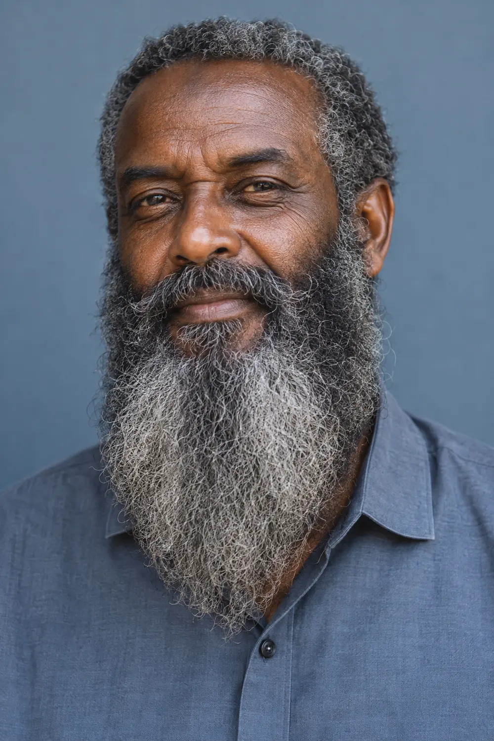 Portrait of an older East African man with a full, wide long beard in coily gray-and-black hair, a connected mustache, and a blue-gray shirt.