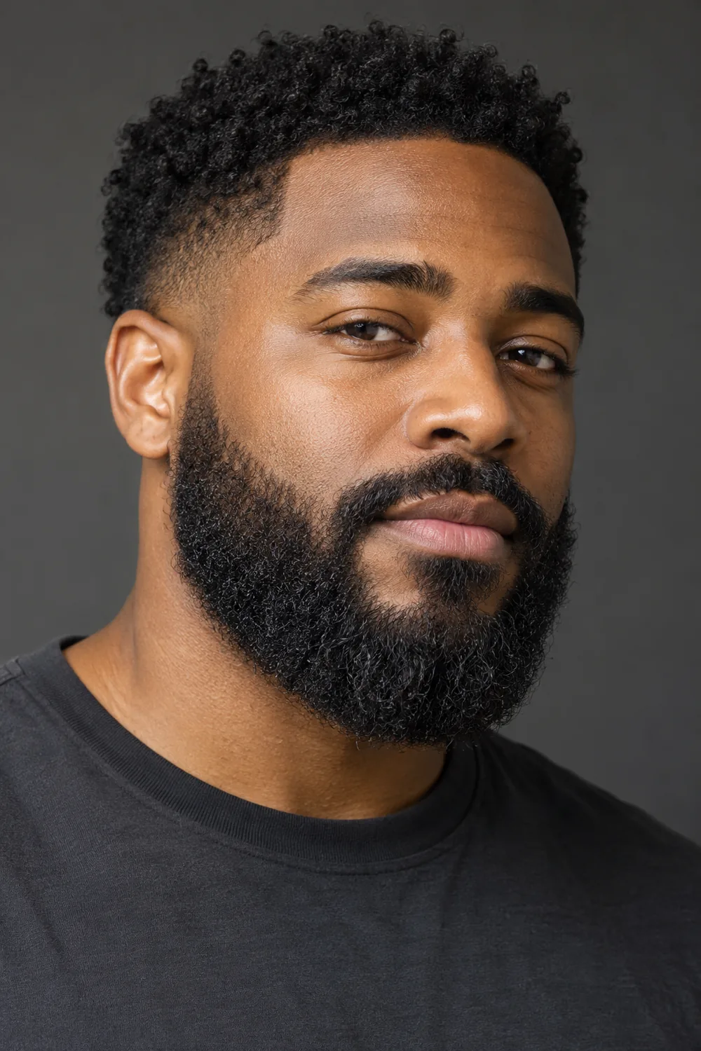 Three-quarter portrait of an adult Black man with a dense, rounded medium-length beard, softly blended cheek edges, tapered sideburns and a connected mustache.