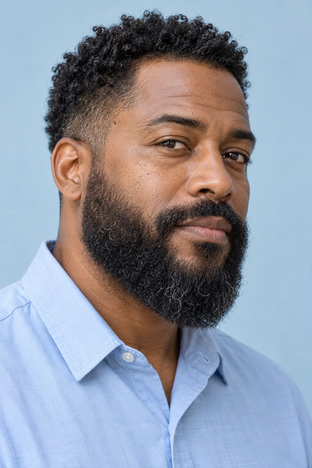 Adult man with a dense, coily medium-length beard, low natural cheek line, gently curved chin and connected mustache, wearing a light blue shirt.