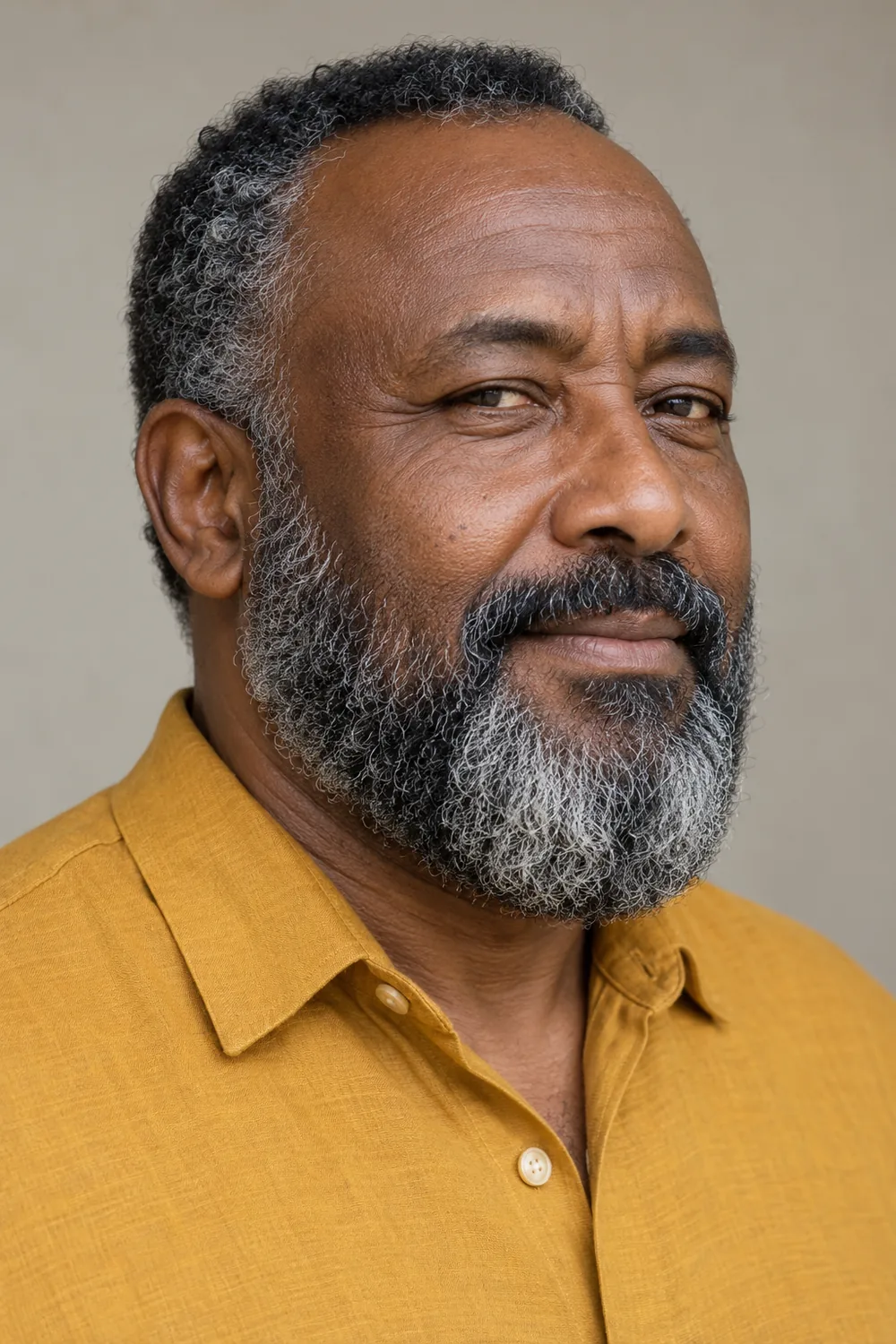 Three-quarter portrait of an adult East African man in a mustard shirt, showing a rounded black, gray, and white beard with a connected mustache and clean neckline.