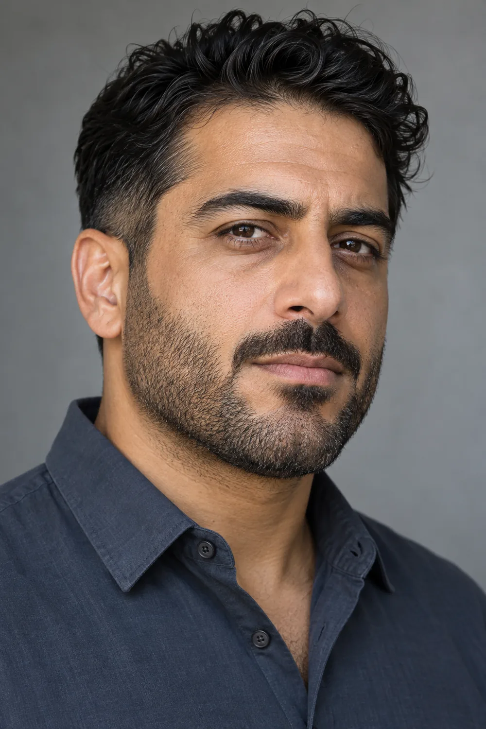Three-quarter portrait of a fictional adult Middle Eastern man with olive-brown skin, heavy short stubble, and a mustache subtly separated from the beard at the corners.