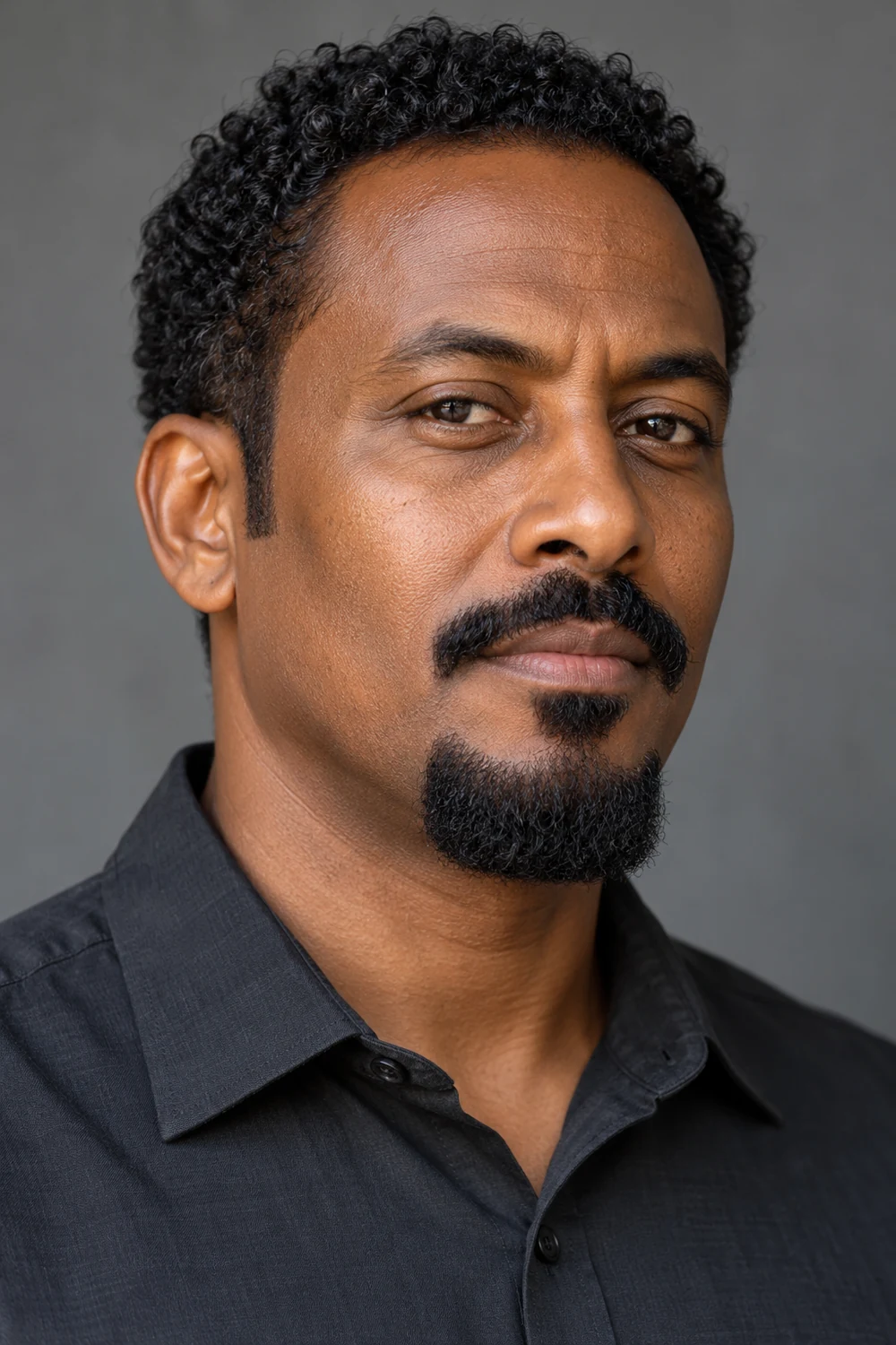 Three-quarter portrait of an adult East African man with tightly curled black hair, a square-bottomed chin beard, disconnected medium mustache, and clean-shaven cheeks.