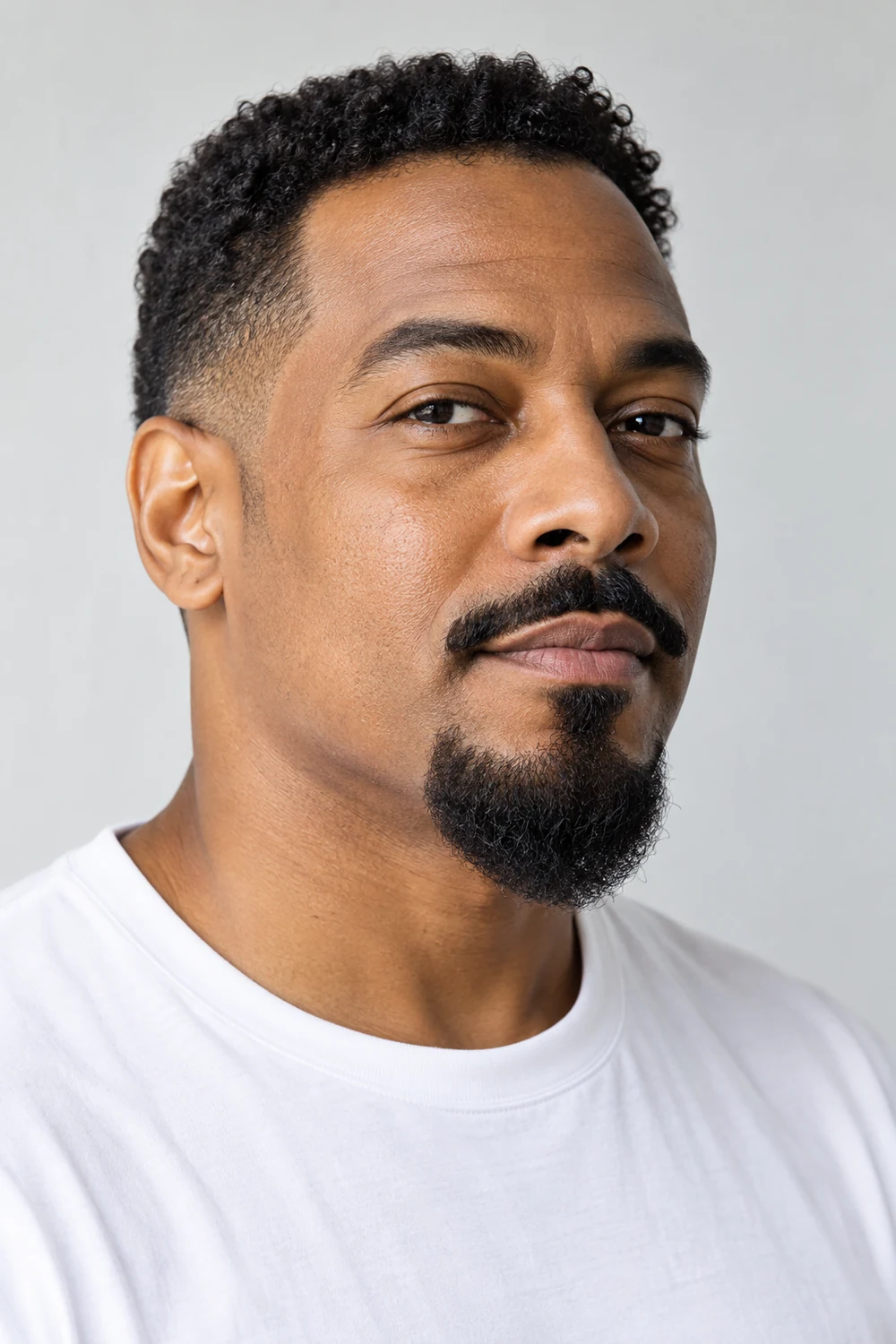 Adult Black man in a three-quarter portrait with a shaped mustache disconnected from a compact, softly pointed curly chin beard and clean-shaven cheeks.