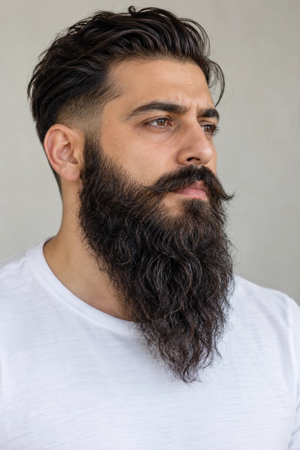 Three-quarter portrait of an adult man with a long, dark wavy beard, tapered point, faded sideburn transition, and connected mustache, wearing a white crew-neck shirt.